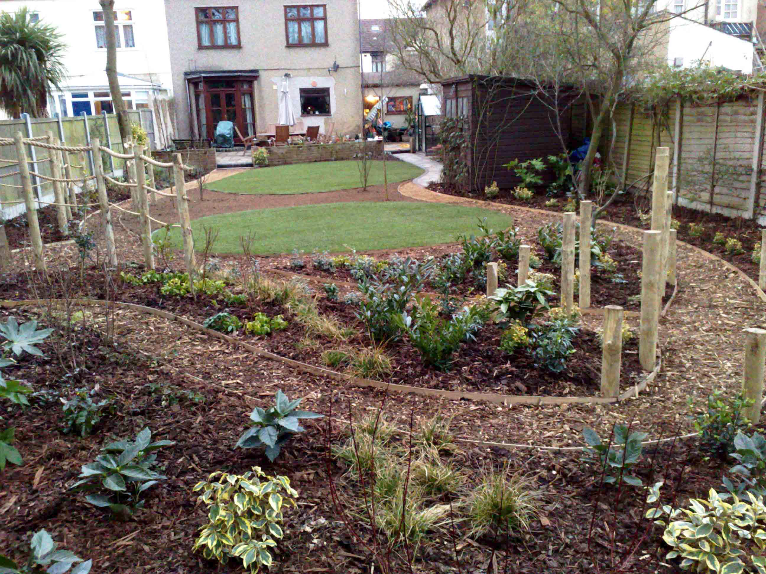 View of garden looking back to the house across the winding shady walk path with shrubs and softwood post crossroad feature in midgorund